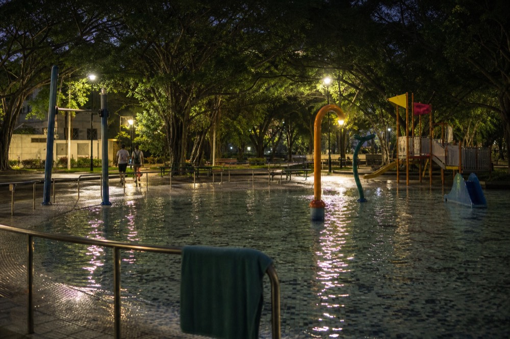 A lit playground and shallow pool area in a city park in the evening, no text, no logos, no people